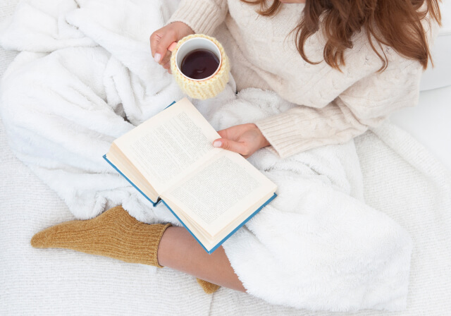 ベッドに座って本を読みながらコーヒーを飲む女性
