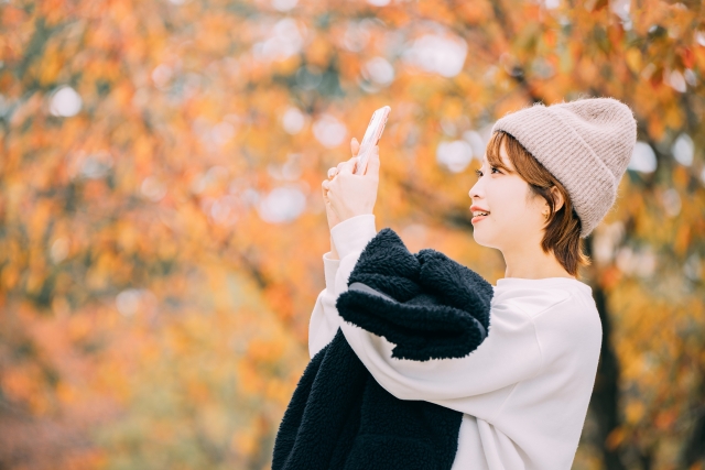 秋の紅葉をスマホで撮影するニット帽の女性