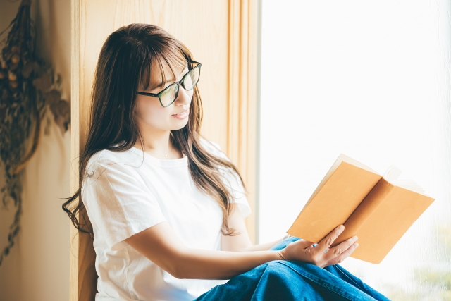 雨の日、窓際に座って読書をする眼鏡の女性